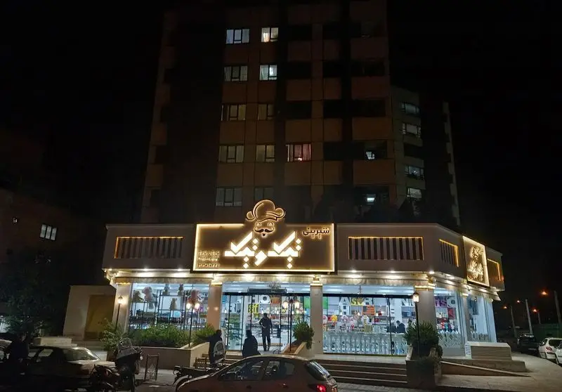 نمای شبانه قنادی تیک و تیک با تابلوی درخشان لوگوی سرآشپز و سبیل. ویترین‌های جذاب شیرینی و حضور مشتریان در ورودی.
