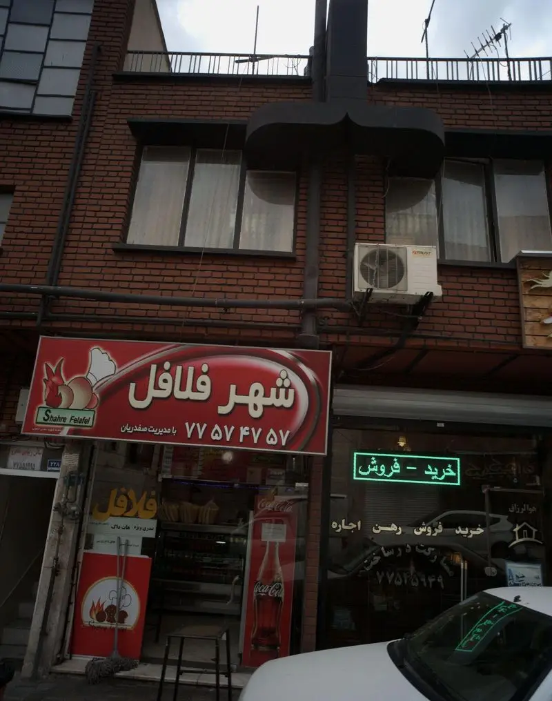 تصویر نمای بیرونی مغازه شهر فلافل، فست فودی معروف با تابلوی قرمز و درخشان. شماره تماس: ۷۷۵۷۴۷۵۷. برای سفارش فلافل و ساندویچ.