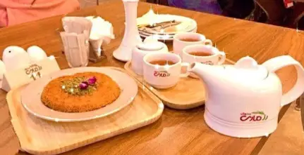 میز چوبی با یک دیس کنافه خوش‌رنگ تزئین شده با پسته و غنچه گل، به همراه قوری و فنجان‌های چای برند رزماری در رستوران رزماری.