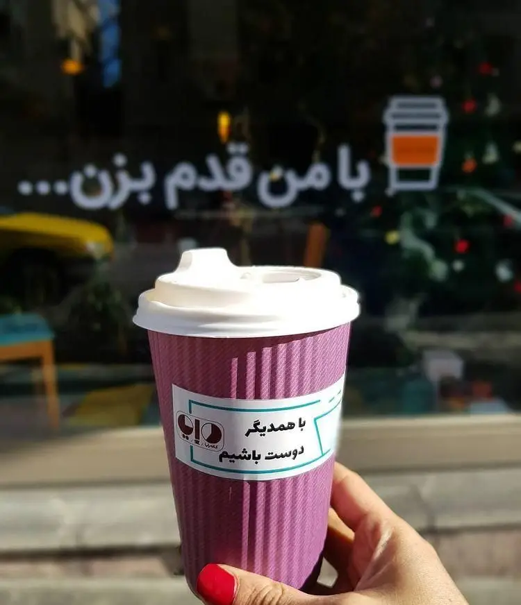 دست لاک‌زده لیوان قهوه بنفش خانه رایا با پیام "با همدیگر دوست باشیم" را نگه داشته است. شعار "با من قدم بزن" در ویترین.