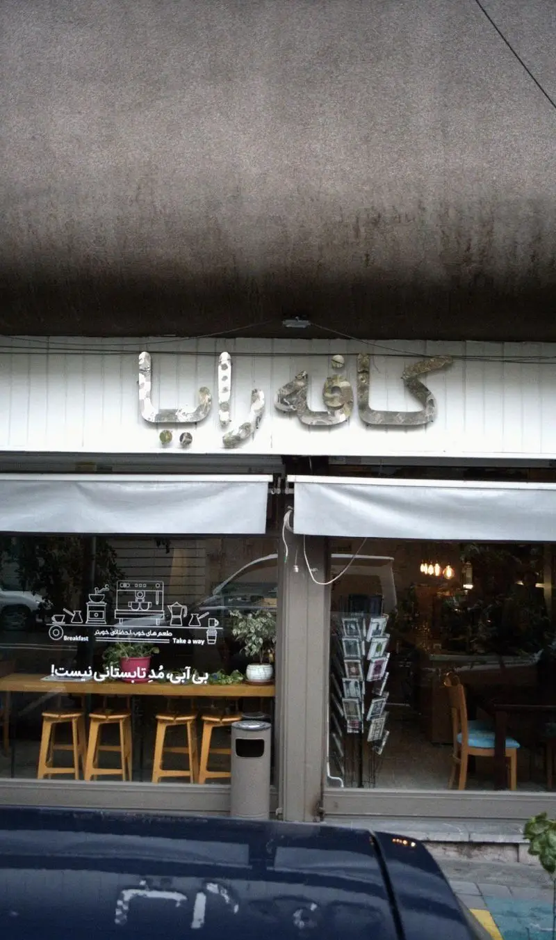 نمای بیرونی کافه خانه رایا، با تابلوی بزرگ و شیشه‌های ویترین که استیکرهای صبحانه و بیرون‌بر دارد. داخل کافه، صندلی‌های چوبی دیده می‌شوند.