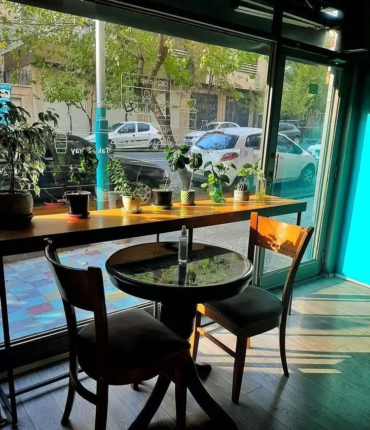 فضای داخلی دنج و پرنور کافه خانه رایا با میز و صندلی چوبی و گلدان‌های سبز کنار پنجره بزرگ رو به خیابان شهر.