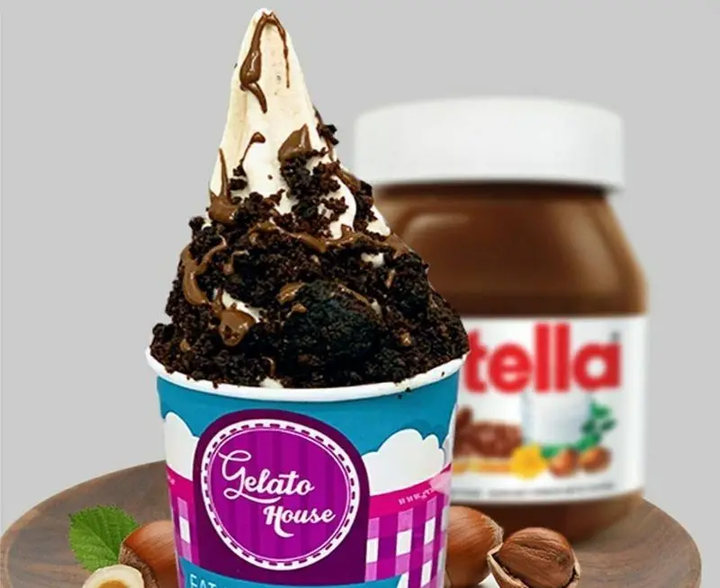 Gelato House: لیوان جلاتو وانیلی با خرده‌های شکلات، سس شکلات و فندق، با شیشه نوتلا در پس‌زمینه. دسر خوشمزه شکلاتی.