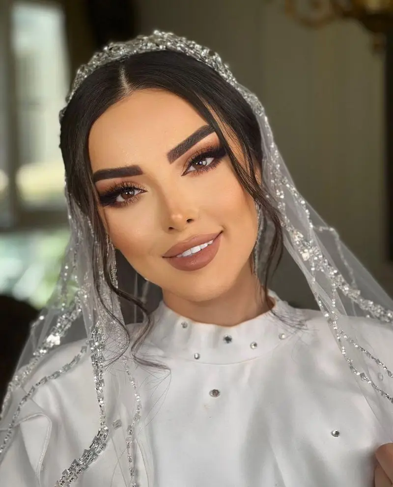تصویر مدل میکاپ و آرایش عروس با رژ لب نود، سایه چشم تیره و تور عروس کار شده با نگین‌های درخشان در سالن زیبایی موزا