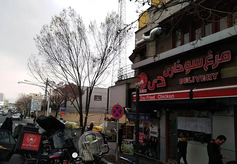 نمای بیرونی رستوران فست فود مرغ سوخاری دی با تابلوهای قرمز، موتورسیکلت‌های دلیوری و فضای خیابان شلوغ شهری.