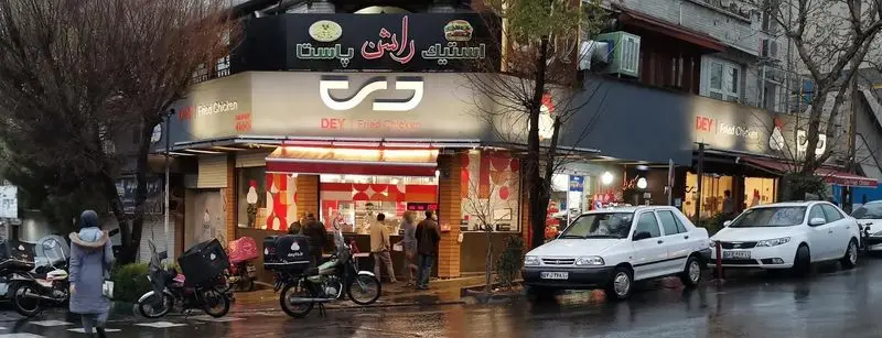 تصویری از نمای خارجی رستوران مرغ سوخاری دی با تابلوی بزرگ و روشن آن، در خیابان شلوغ با مردم، خودروها و موتورسیکلت‌های پیک.