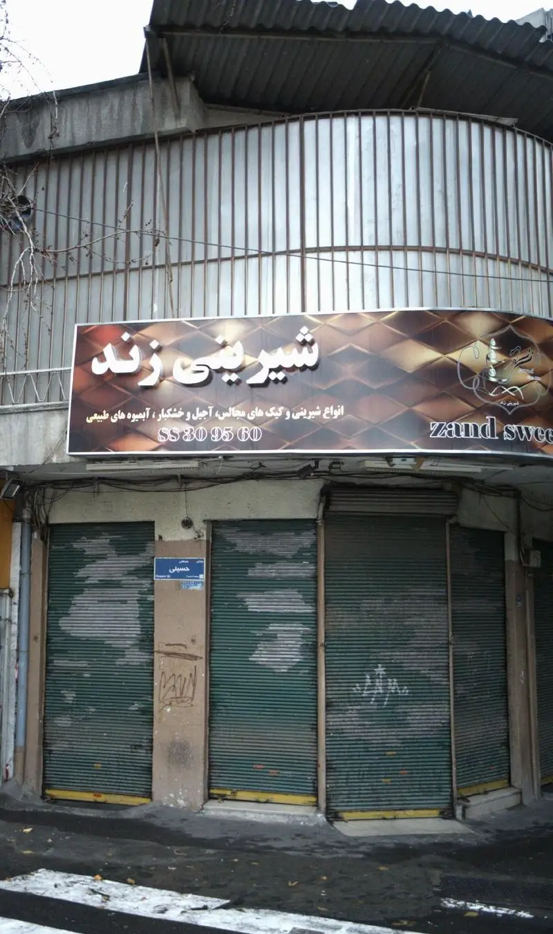 نمای بیرونی مغازه شیرینی زند در خیابان حسینی با کرکره‌های بسته و تابلوی خدمات شامل انواع شیرینی، آجیل و آبمیوه طبیعی.