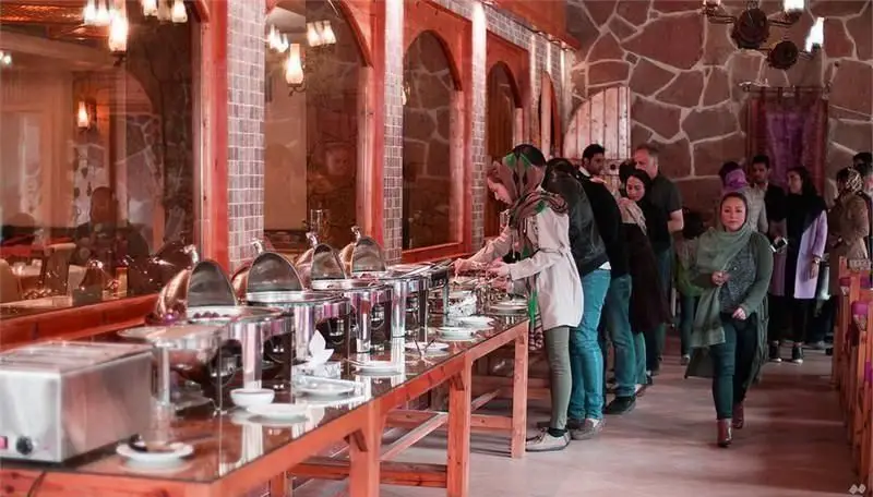 صف بوفه غذای پرطرفدار رستوران قصر موج، مشتریان در حال انتخاب غذاهای لذیذ در فضایی سنتی با معماری سنگی و نورپردازی گرم و دلپذیر.