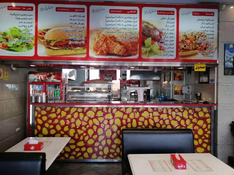 تصویری از داخل فست فود دلفین، شامل منوهای متنوع پیتزا، برگر، ساندویچ، کنتاکی و پیش غذا، همراه با میز و صندلی‌ها و پیشخوان تمیز رستوران.