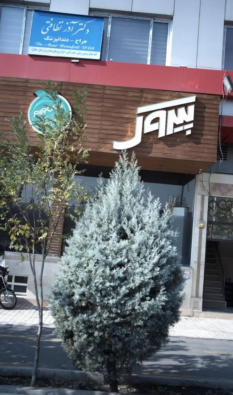 نمای کلی از پیتزا پیروک در یک روز آفتابی، شامل تابلوی برند با حروف سفید و نمای چوبی ساختمان با درختان.