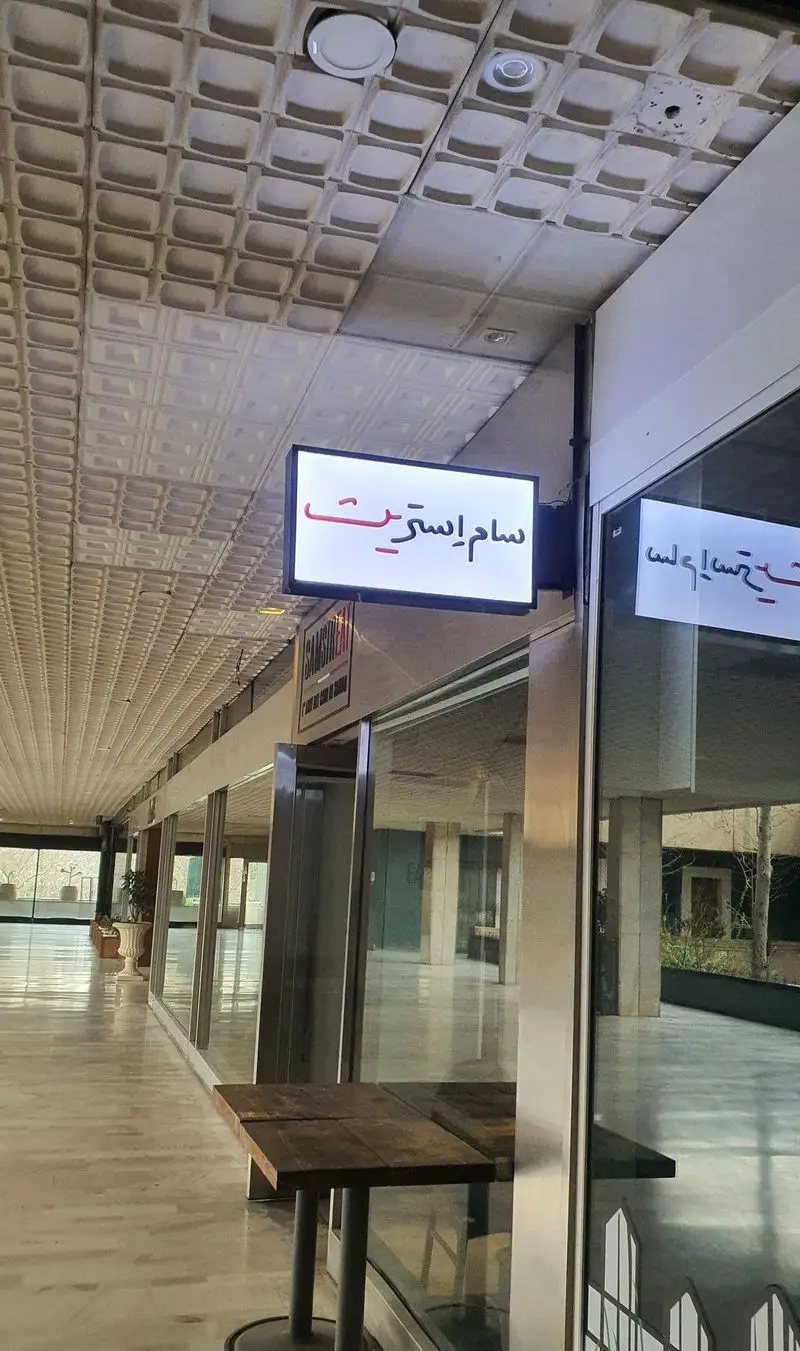 فست فود سام استریت، تابلوی روشن در ورودی یک راهروی وسیع با سقف مربعی، میز چوبی در جلوی ویترین، فضایی شیک و مدرن.