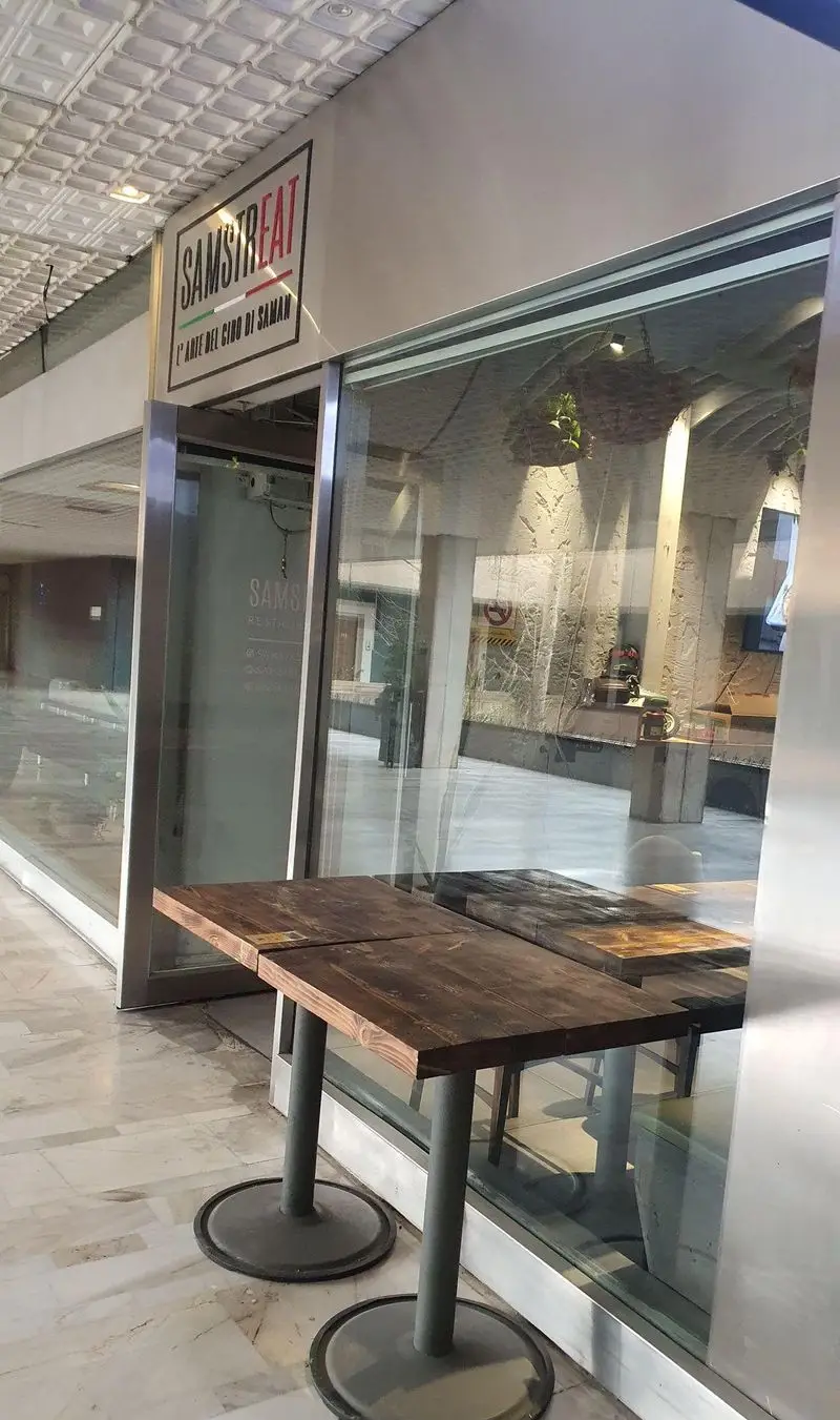 نمای بیرونی فست فود و رستوران سام استریت (SAMSTREAT) با میزهای چوبی جلوی ورودی. تابلوی خاص آن با طرح پرچم ایتالیا.