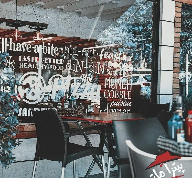 نمای بیرونی و فضای باز پیتزا هات پاسداران با دکوراسیون مدرن و جملات غذایی روی شیشه، محیطی دلنشین برای صرف پیتزا و غذا ارائه می‌دهد.