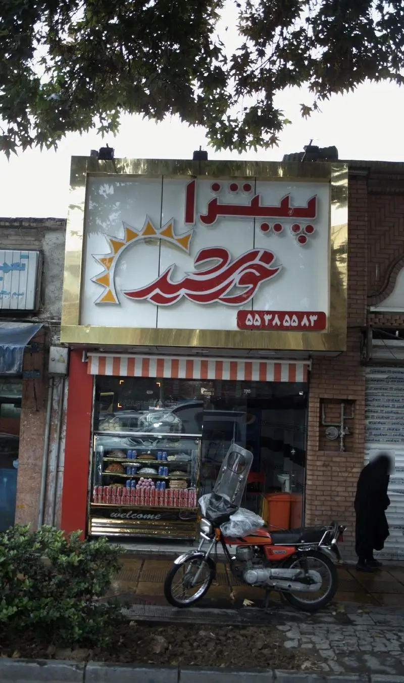 تابلوی درخشان فست فود پیتزا خورشید با لوگوی خورشید و شماره ۵۵۲۸۵۵۸۳، ویترین جذاب خوراکی‌ها و موتور پارک شده در نمای بیرونی.