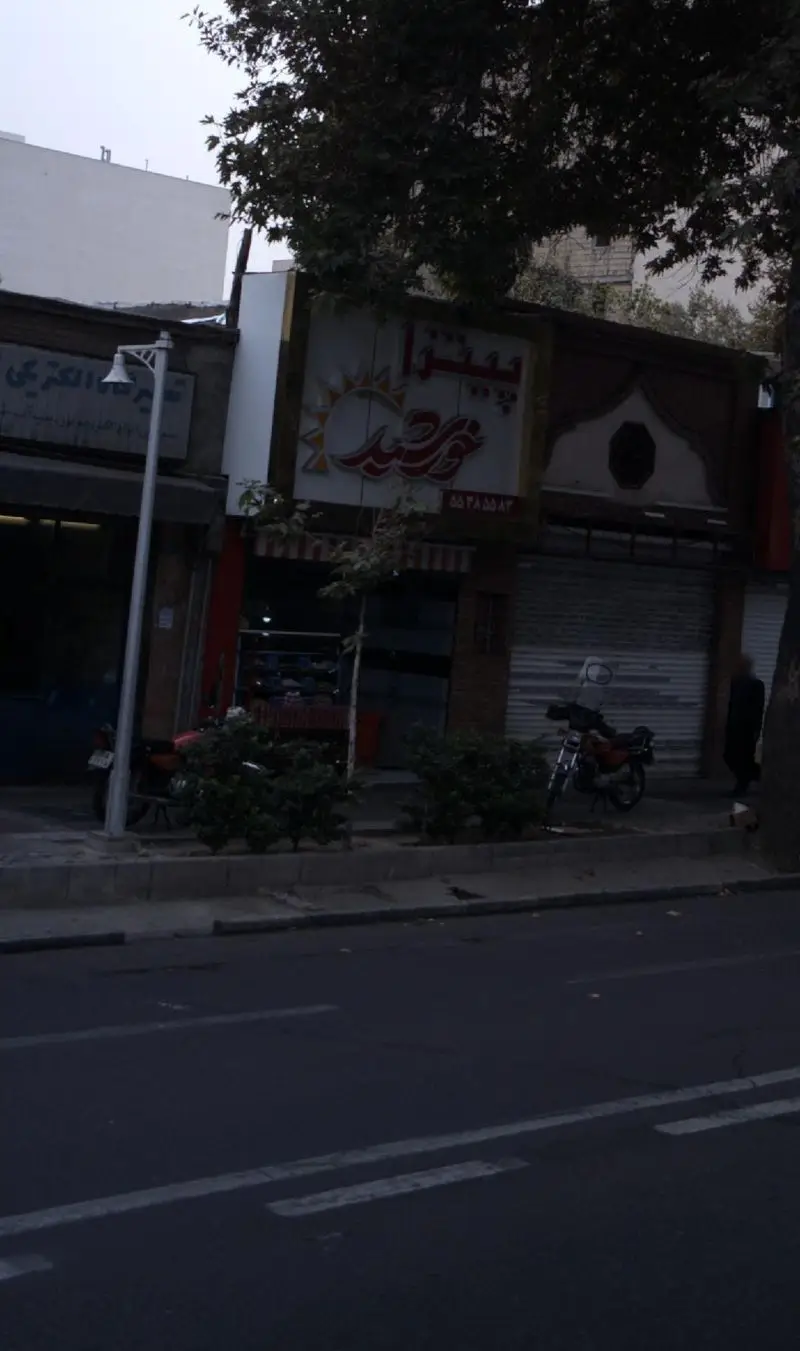 تصویری از نمای بیرونی مغازه فست فود پیتزا خورشید در خیابان، با تابلوی نام و لوگوی خورشید، در هوای گرگ و میش.