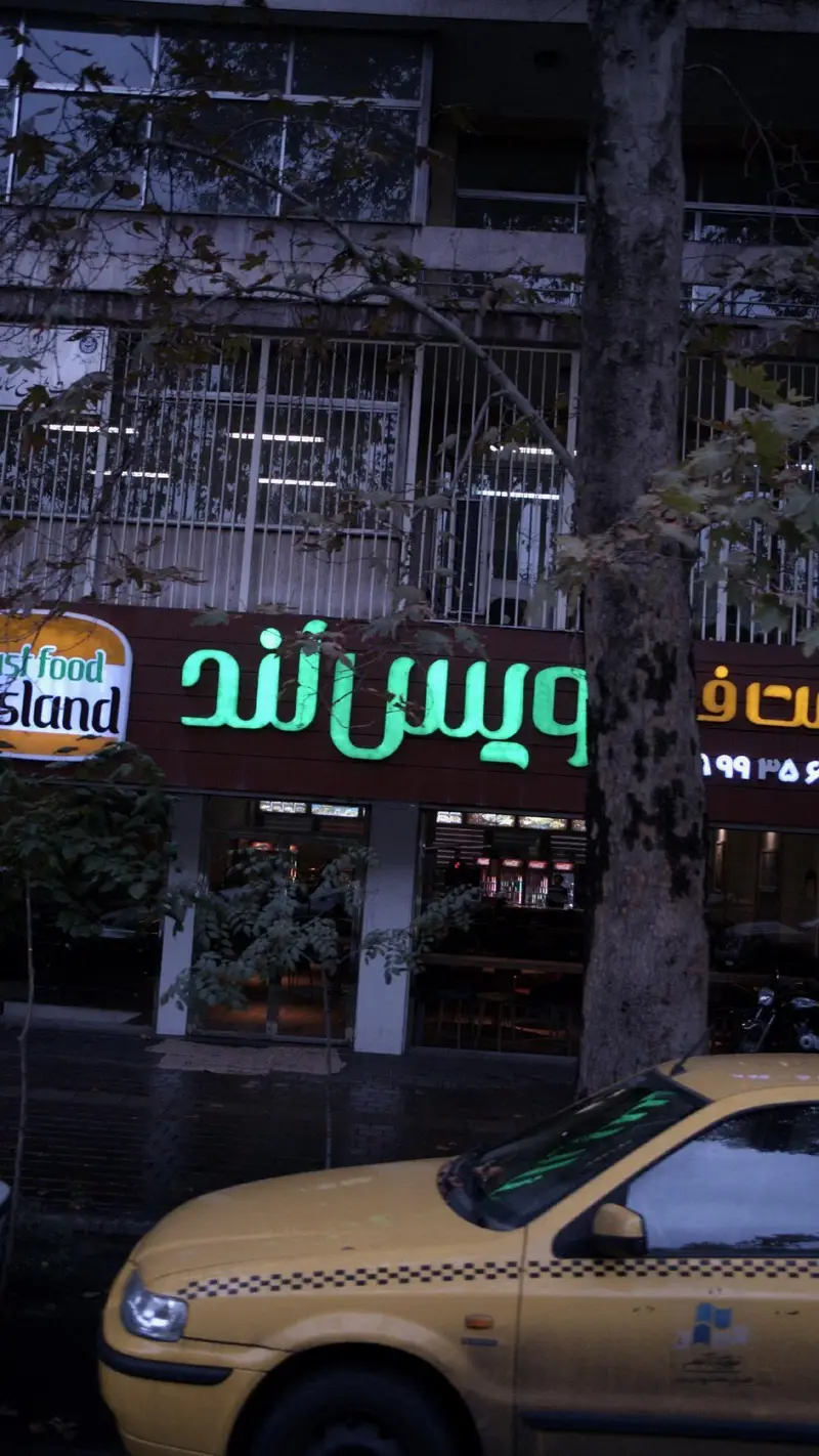 فست فود ویسلند با تابلوی نئون سبز روشن و نام تجاری "Fast Food Island" در شب. نمای خیابان با تاکسی زرد و درختان.