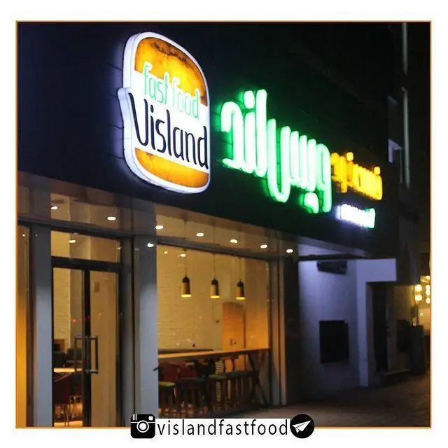 تابلوی روشن فست فود ویسلند در شب؛ لوگوی همبرگر و نام فروشگاه به فارسی و انگلیسی. نمای خارجی رستوران مدرن با ویترین شیشه‌ای.