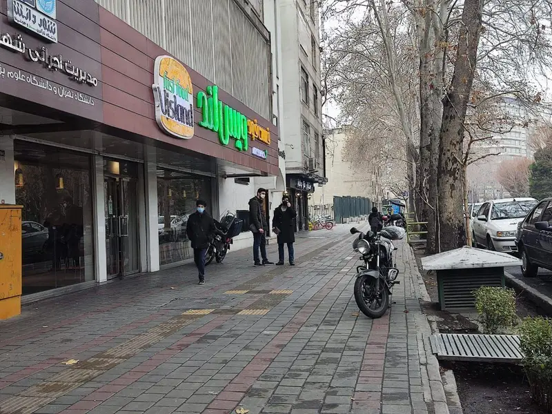 نمای بیرونی فست فود ویس لند در یک روز ابری، تابلوی روشن رستوران، پیاده‌رو با عابران ماسک‌زده و موتورسیکلت پارک شده.