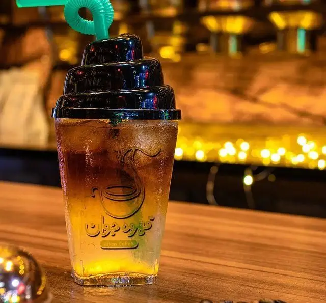 یک لیوان آیس کافی لذیذ کافه جوان با لایه‌های قهوه و نوشیدنی، نی سبز و درب فانتزی در فضایی دلنشین