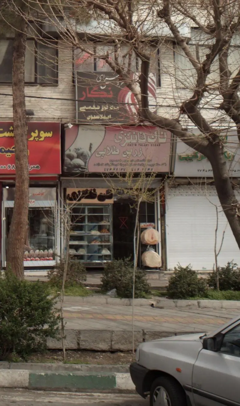 نمای بیرونی فروشگاه نان فانتزی زرین طلایی با تابلوی بزرگ آن، ویترین پر از نان و شیرینی، در کنار خیابان، درختان بدون برگ و خودرویی پارک شده.