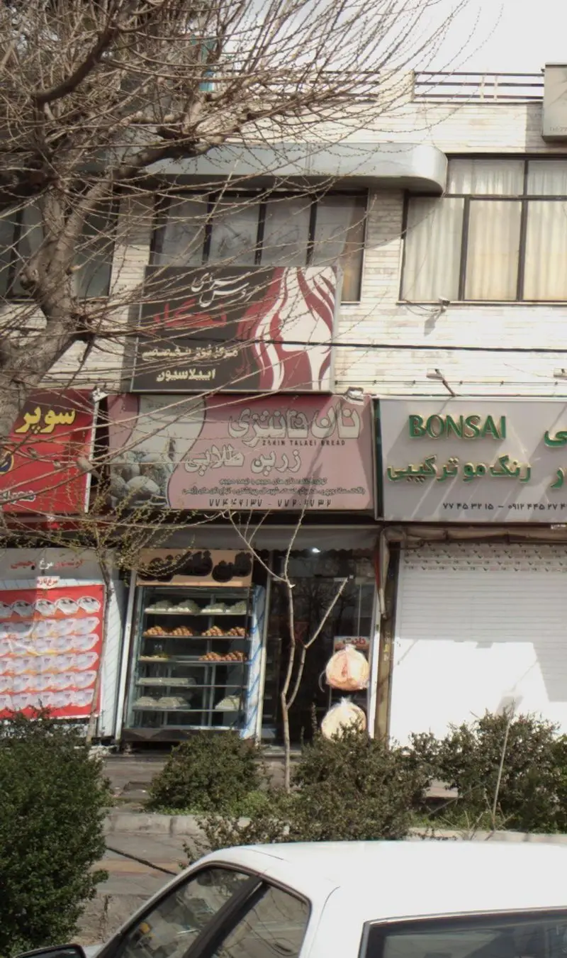 نمای بیرونی مغازه نان فانتزی زرین طلایی با تابلو، محصولات پخت شده و شیرینی‌های تازه در ویترین، در خیابان شلوغ شهری