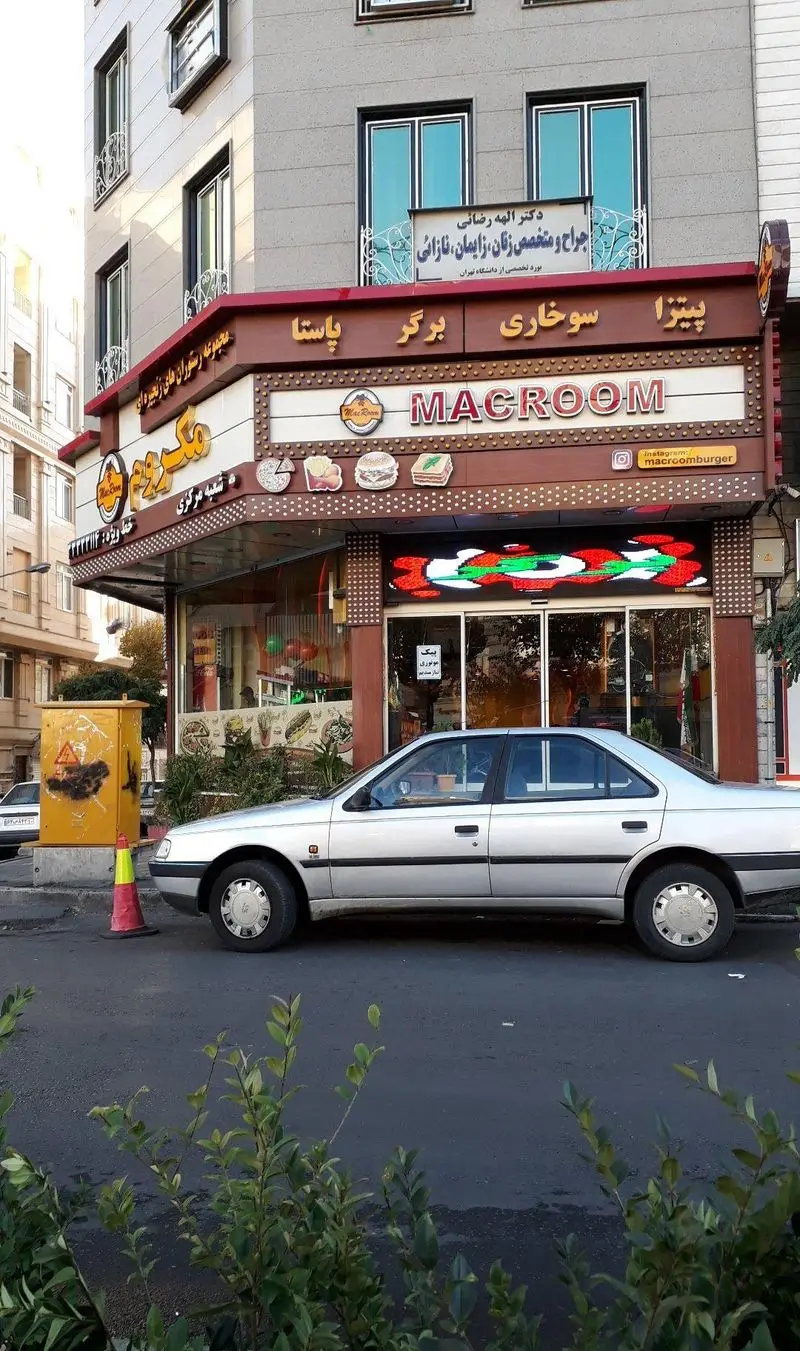 نمایی از رستوران فست فود مکروم (پیتزا، برگر، سوخاری، پاستا) در یک ساختمان گوشه با خودروی نقره ای و تابلوی روشن.