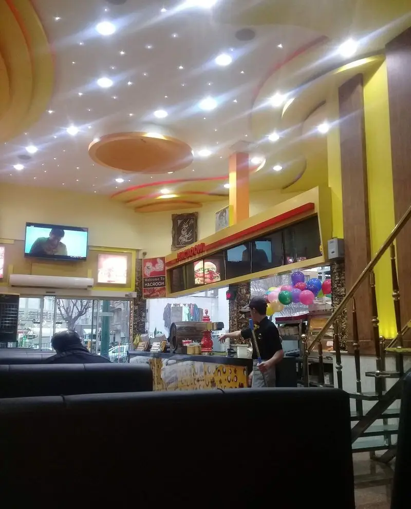 فضای داخلی دنج و مدرن پیتزا مکروم با سقف نورانی، مشتریان نشسته، و پرسنل در حال کار. محیطی عالی برای فست فود و پیتزا.