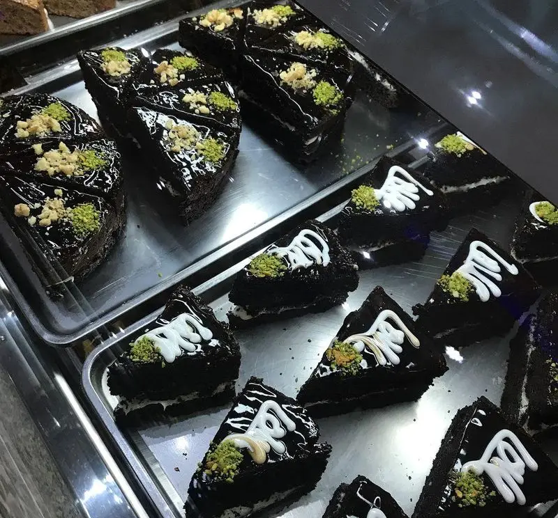 دسرهای شکلاتی تیره و براق کافه قنادی لادن، تزئین شده با پودر پسته، مغز آجیل و طرح‌های آیسینگ سفید. شیرینی‌جات لذیذ.