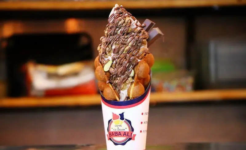 تصویری از بستنی بابا علی با وافل حبابی پر شده از بستنی، روکش سس شکلات، آجیل مخلوط و تکه‌های شکلات، یک دسر لذیذ و پرطرفدار