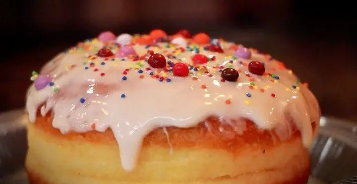 تصویری اشتهاآور از یک دسر دونات یا شیرینی بزرگ با تزیینات رنگی و لعاب سفید، در منوی خاص بستنی بابا علی.