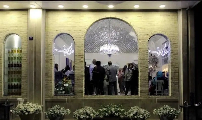 نمای شیک رستوران دلی جان در شب با پنجره‌های قوسی، معماری آجری و تابلوی نورانی "دلی جان" به خط نستعلیق. فضای داخلی پر رفت‌وآمد.