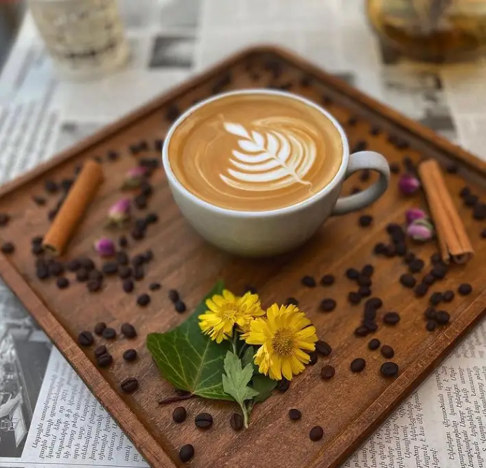 فنجان لاته با طرح برگ روی سینی چوبی، تزئین شده با دانه‌های قهوه، چوب دارچین و گل‌های زرد در محیط دلنشین کافی شاپ فرندز.