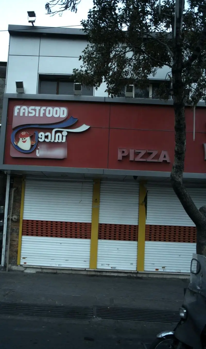 تصویر تابلوی فست فود و پیتزا نمکدون با زمینه قرمز، شامل کلمات FASTFOOD و PIZZA و لوگوی خاص آن، نمای جلوی رستوران.