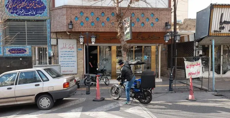 تصویر خیابانی از نمای بیرونی کترینگ کاظمی در یک روز آفتابی. موتورسوار در حال آماده شدن برای تحویل سفارشات غذایی.