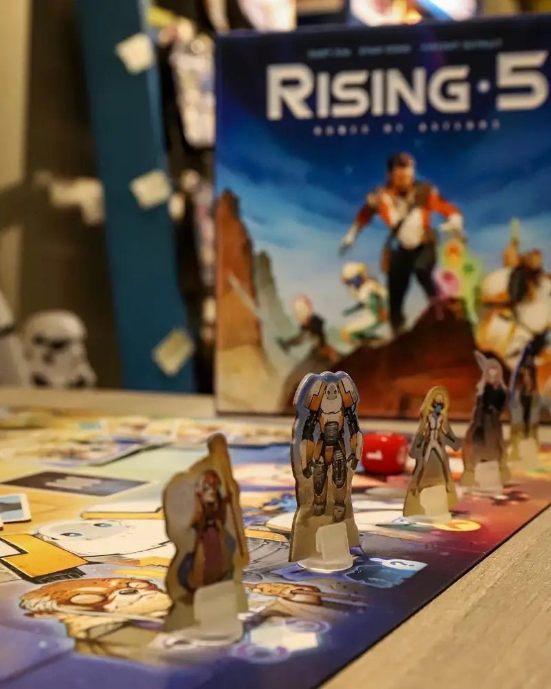 نمایی نزدیک از بازی رومیزی Rising 5 روی میز کافه برد، شامل شخصیت‌های کارتونی ایستاده، تاس و جعبه بازی اصلی. محیطی جذاب برای گیمرها.