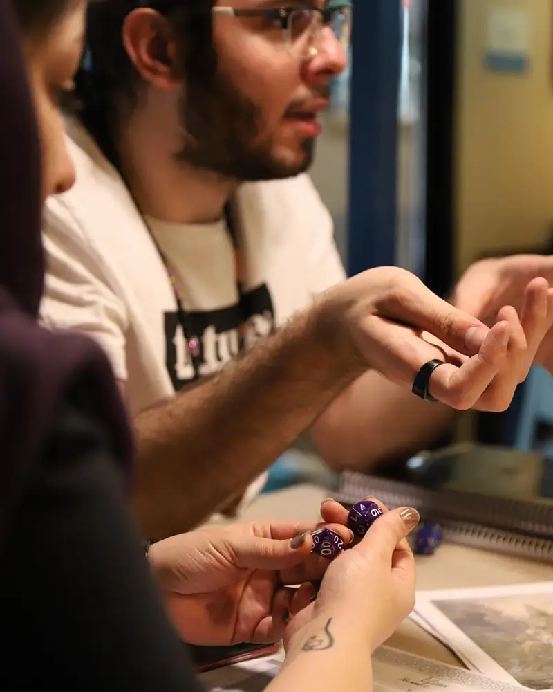 تصویری از دو نفر در کافه برد، غرق در یک بازی رومیزی نقش‌آفرینی هیجان‌انگیز با تاس‌های ۲۰ وجهی بنفش. محیطی دوستانه و پرنشاط.