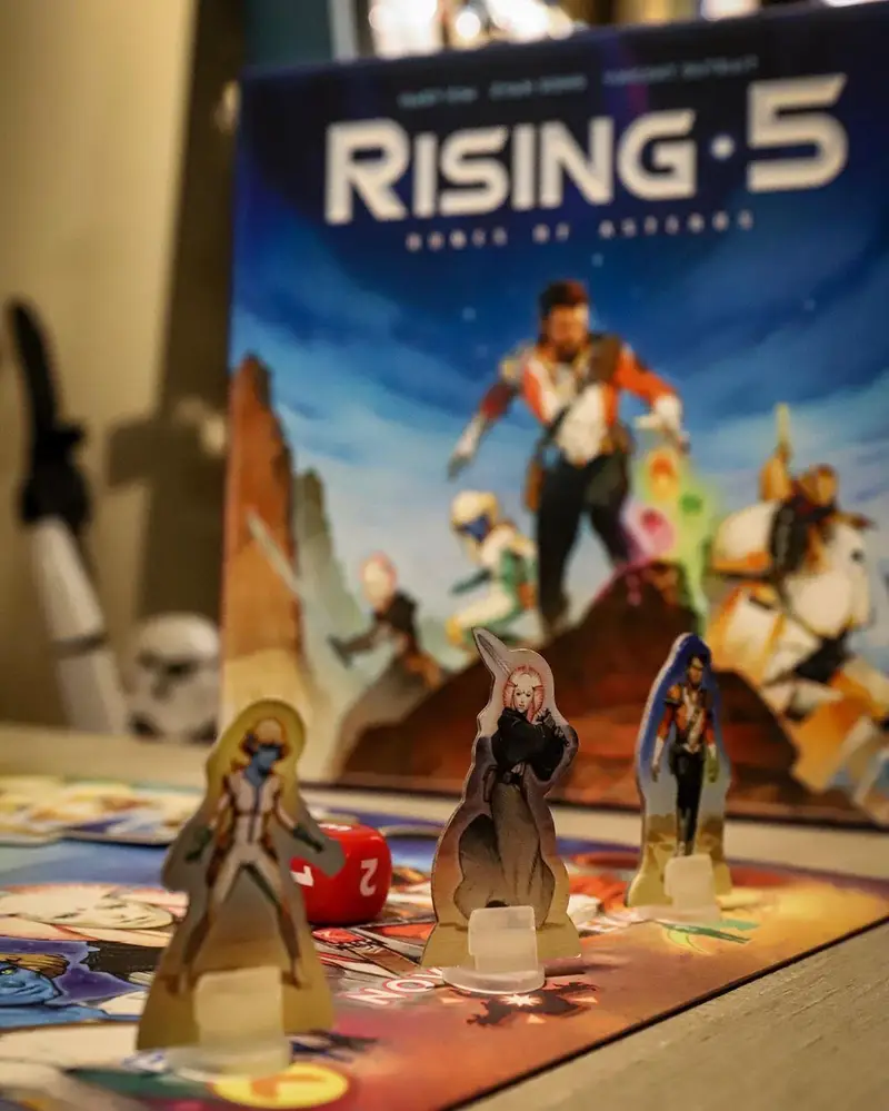 نمایی از بازی رومیزی Rising 5: Runes of Asteros در کافه برد، با فیگورهای شخصیت‌ها، صفحه بازی و تاس. لحظاتی پرهیجان در یک بازی فکری.