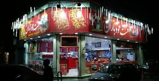 نمای شبانه و جذاب فست فود باران، رستورانی با تابلوی قرمز روشن، پیتزا، همبرگر و فضای داخلی پر از مشتریان در حال غذا خوردن.