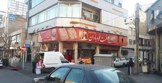 فست فود باران، رستورانی با تابلوی قرمز و جذاب، ارائه دهنده انواع پیتزا و برگر، در نمای خیابان با اتومبیل‌ها و مردم.