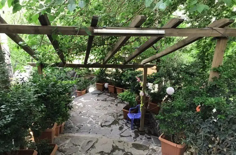 فضای سبز و دلنشین رستوران ایرانی آرامیس با آلاچیق چوبی، پله‌های سنگی و گلدان‌های گل، مکانی آرام برای صرف غذا در طبیعت.