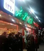 نمای شبانه از ویتامینه و بستنی ایوب با جمعیت مشتریان، تابلوهای نئونی درخشان و سال تاسیس ۱۳۴۰. مکانی پرجنب‌وجوش برای دسر و نوشیدنی.