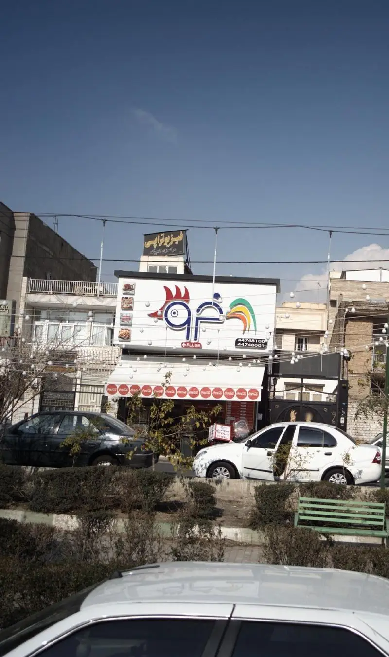 تصویر نمای خارجی فست فود پلاس خروس (+PLUSE)، تابلوی بزرگ با لوگوی خروس رنگین‌کمانی، منوی غذا و شماره تماس: 44748100.