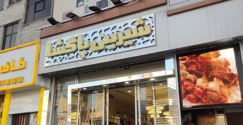 تصویر نمای بیرونی فروشگاه شیرینی و قنادی پاگشا با تابلوی روشن و ویترین شیشه ای نمایشگر انواع شیرینی ها و کیک های خوشمزه.