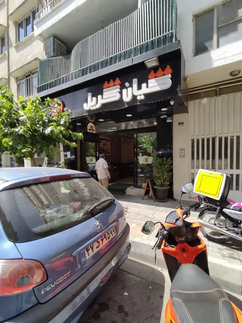 نمای بیرونی مغازه فست فود کیان گریل با تابلوی بزرگ و مشخص، همراه با خودروی آبی و موتور سیکلت‌های پارک شده در جلوی ورودی.