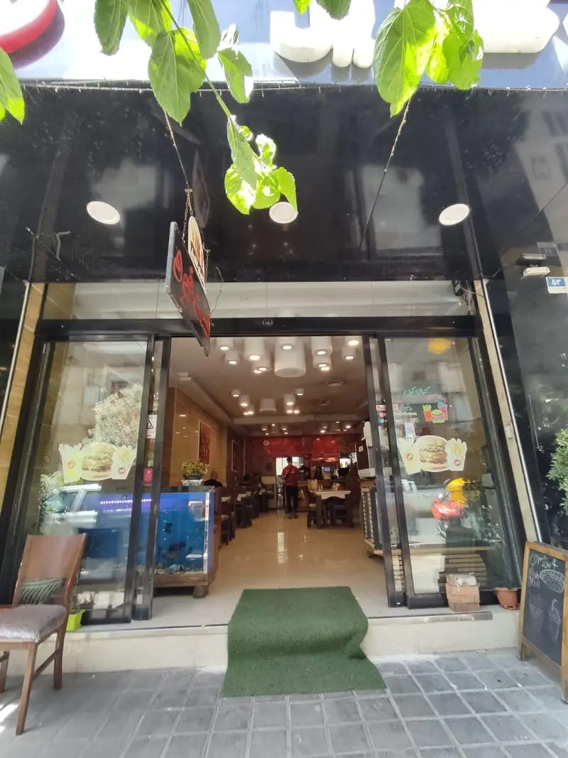 نمای خارجی فست فود کیان گریل با تابلوی سفید روی زمینه مشکی، شاخه‌های سبز و ورودی شیشه‌ای باز به داخل رستوران مدرن و پرنور.