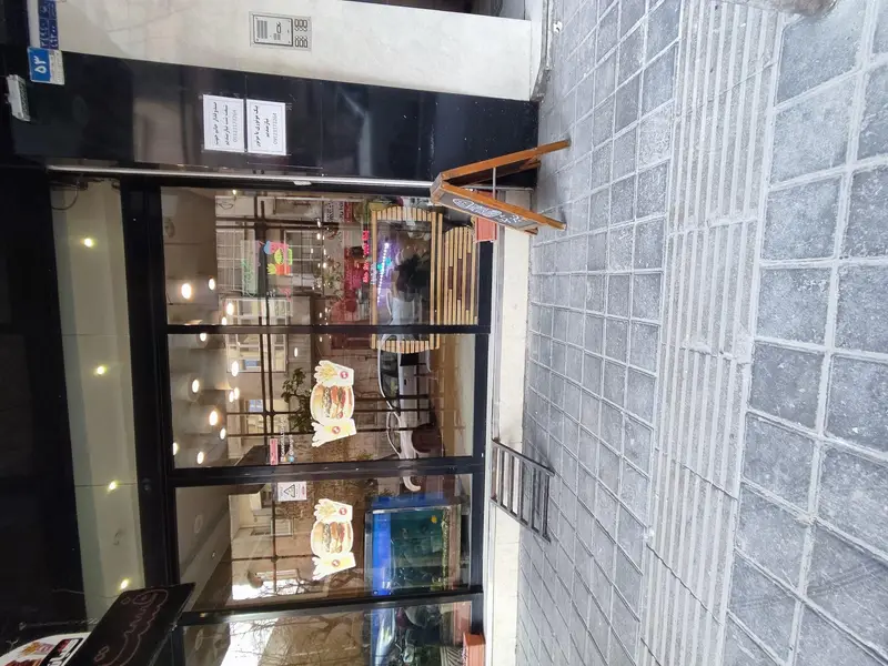 ورودی اصلی فست فود کیان گریل با درب‌های شیشه‌ای بزرگ، برچسب همبرگر روی ویترین، نمای سیاه رنگ و پیاده‌رو طوسی سنگفرش شده با تابلوی A-frame.