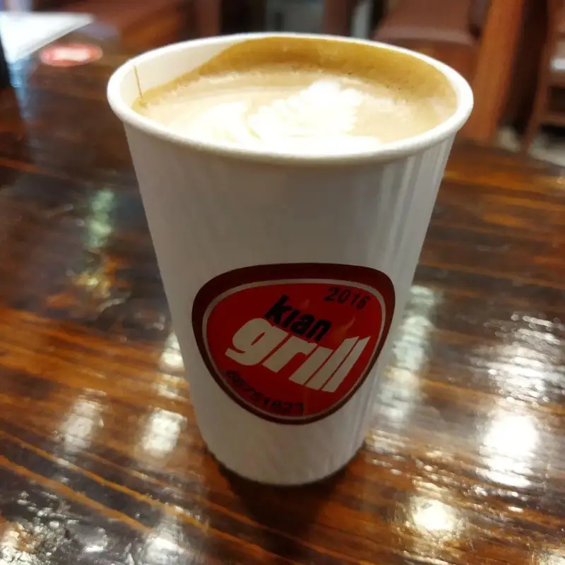 تصویری از یک لیوان نوشیدنی گرم لاته با فوم آرت و لوگوی برجسته فست فود کیان گریل Kian Grill روی میز چوبی رستوران.