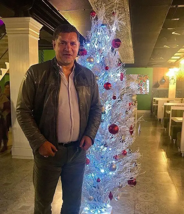 مردی خوش‌پوش در رستوران کمالی کنار درخت کریسمس سفید و نورانی، نشان‌دهنده فضای گرم و دکوراسیون جذاب برای مشتریان.