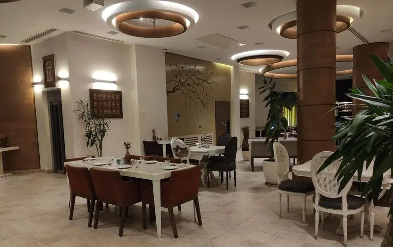 فضای داخلی مدرن و دلباز رستوران کمالی با میزهای غذاخوری چیده شده، صندلی‌های راحت، نورپردازی شیک و دیوارنگاره درخت.