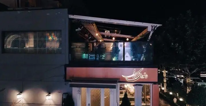 رستوران کمالی در شب، نمای بیرونی با تابلوی روشن، فضای باز روف گاردن مدرن با چوب و نورپردازی زیبا، مناسب برای شام و دورهمی.